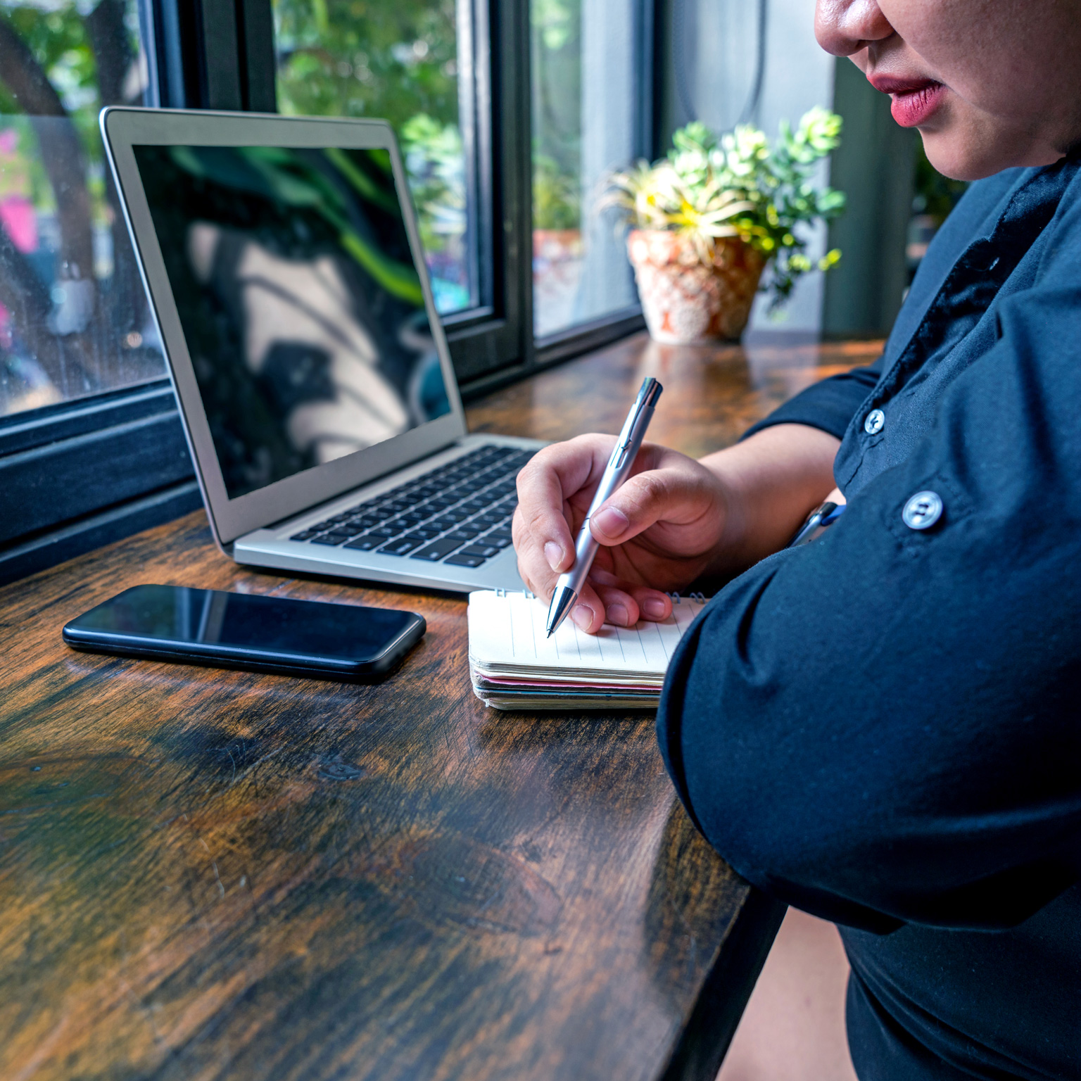 Woman in denim shirt, cropped with her mostly off to the side of the image, with laptop and smartphone, writing in a notebook at a wooden table