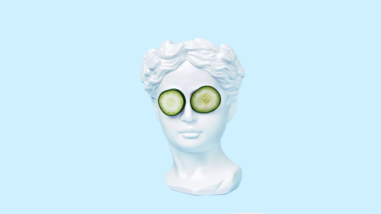 A bust of a young woman, inspired by ancient Greek art, wearing cucumber slices on her eyes and set against a light blue backdrop.