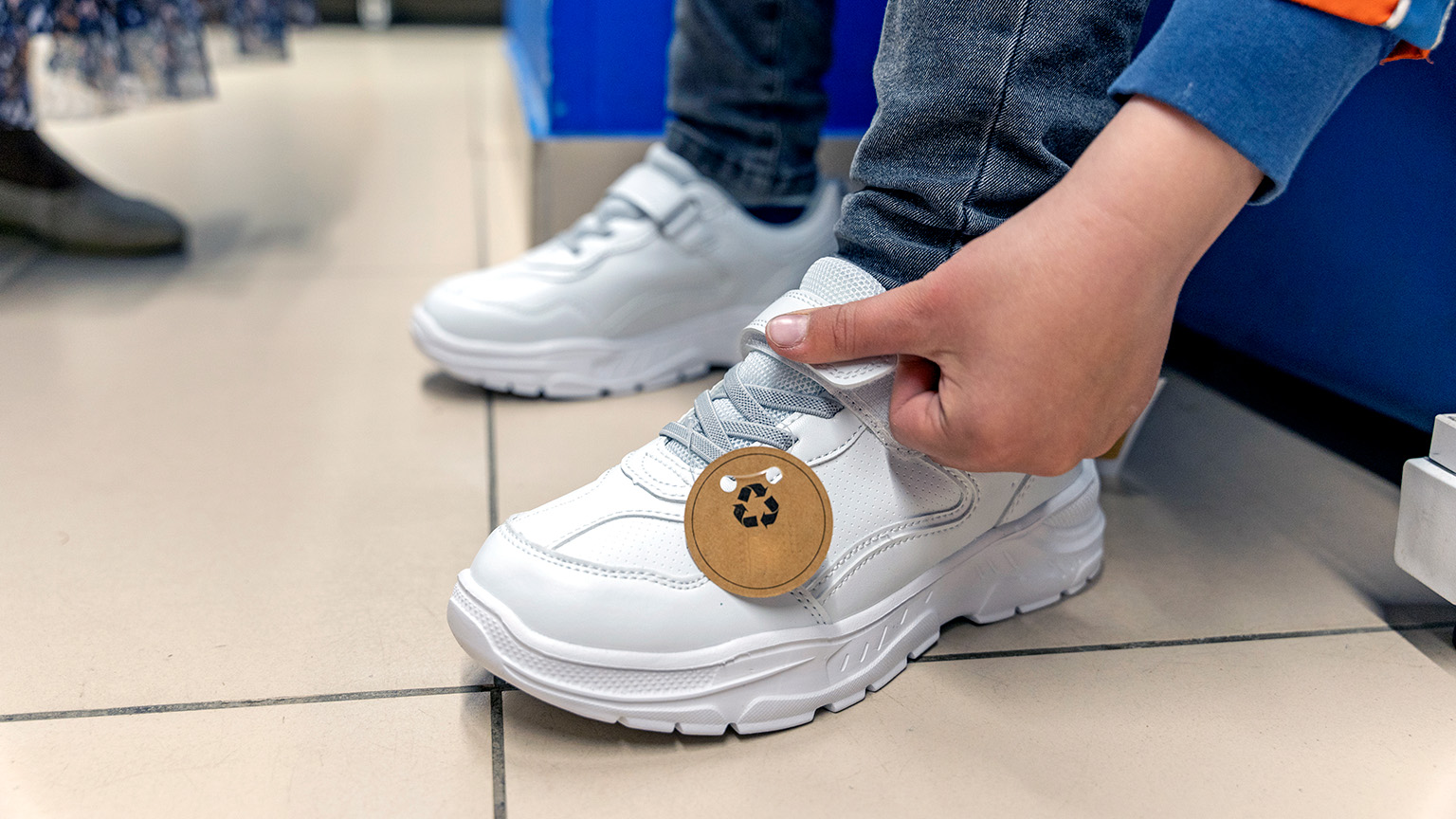 A young boy trying on a new pair of sneakers in a store, with a noticeable recycled tag attached to one of the shoes.